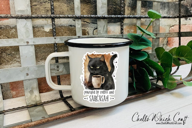Puede incluir: Taza de esmalte blanco con borde y asa negros. La taza tiene una pegatina de un gato negro sosteniendo una taza de caf&eacute;, con el texto "powered by coffee and sarcasm". Un art&iacute;culo humor&iacute;stico.