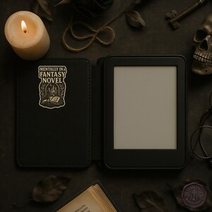May include: Black e-reader with a cover featuring the text "MENTALLY IN A FANTASY NOVEL" and a graphic of a person reading. The scene includes a lit candle, a skull, a key, and other gothic-themed accessories.