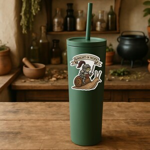 May include: A green tumbler with a straw and a sticker featuring a knight riding a snail, with the text "KNIGHT OF NOPE". The knight is holding a sword and a banner. The background is a wooden surface.