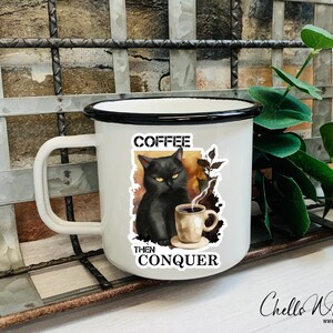 May include: White enamel mug with a black rim and handle. The mug features a black cat with yellow eyes drinking coffee. The text on the mug reads "Coffee Then Conquer".