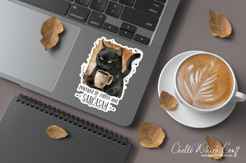 Puede incluir: Una pegatina con un gato negro sosteniendo una taza de caf&eacute;, con el texto "powered by coffee and sarcasm". La pegatina est&aacute; en un port&aacute;til, junto a una taza de caf&eacute; con arte latte y hojas de oto&ntilde;o.