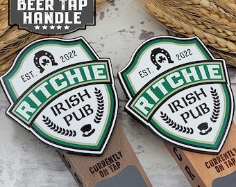 May include: Two personalized beer tap handles with a green, white, and gold Irish pub design. The handles feature the text "EST. 2022 Ritchie Irish Pub" and a shamrock with a horseshoe. The handles are made of wood and have a chalkboard area for writing the name of the beer on tap.