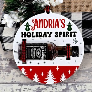 May include: A white, round Christmas ornament with the text "ANDRIA'S HOLIDAY SPIRIT". It features a miniature Jack Daniel's whiskey bottle secured with red and black ribbon. The bottom has a red border with white Christmas trees.