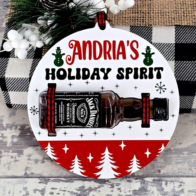 May include: A white, round Christmas ornament with the text "ANDRIA'S HOLIDAY SPIRIT". It features a miniature Jack Daniel's whiskey bottle secured with red and black ribbon. The bottom has a red border with white Christmas trees.