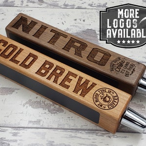 May include: Two wooden beer tap handles with engraved text. The top handle is walnut wood and says "NITRO" with a coffee cup design and the text "Drink your coffee, it's chaos but there". The bottom handle is light wood and says "COLD BREW" with a coffee cup design and the text "My blood type is coffee".