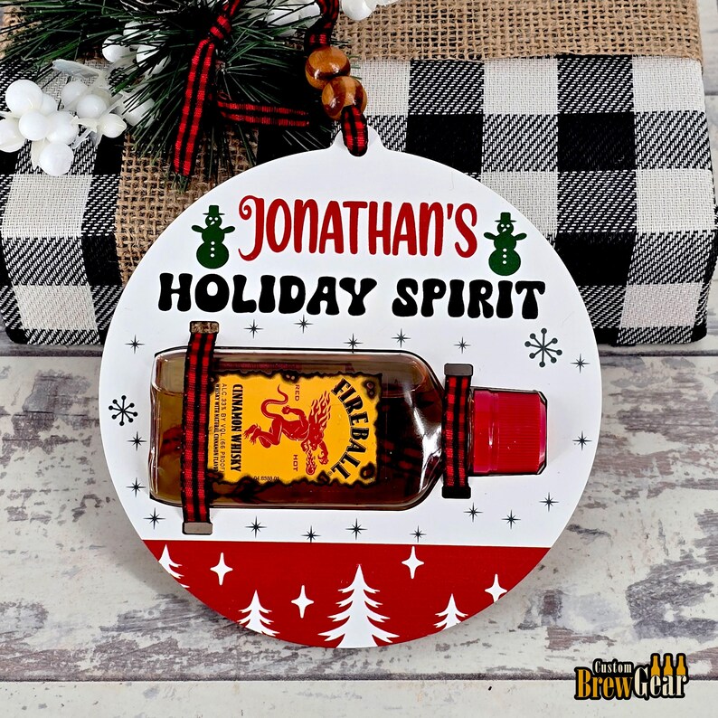 May include: A round, white holiday ornament with the text "Jonathan's Holiday Spirit." It features a small bottle of Fireball Cinnamon Whisky, secured with red and black ribbon. The bottom has a red band with white trees and stars.