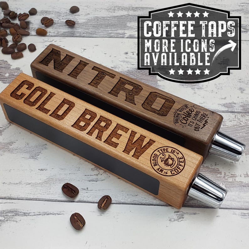 May include: Two wooden beer tap handles, one labeled "NITRO" and the other "COLD BREW". Both handles have a black chalkboard surface and a chrome tap spout. The handles are on a white wooden surface with coffee beans scattered around.