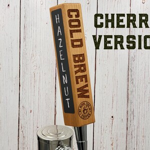 May include: A wooden beer tap handle with the words "COLD BREW" and "HAZELNUT" written on it. The handle also has a logo that says "My Blood Type is Coffee". The text "CHERRY VERSION" is written in green letters on a white wooden background.