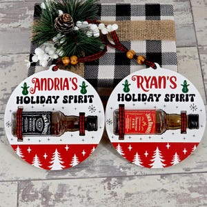 May include: Two round Christmas ornaments with the text "Andria's Holiday Spirit" and "Ryan's Holiday Spirit". Each ornament holds a miniature Jack Daniel's whiskey bottle. The ornaments have a red and white Christmas tree design.