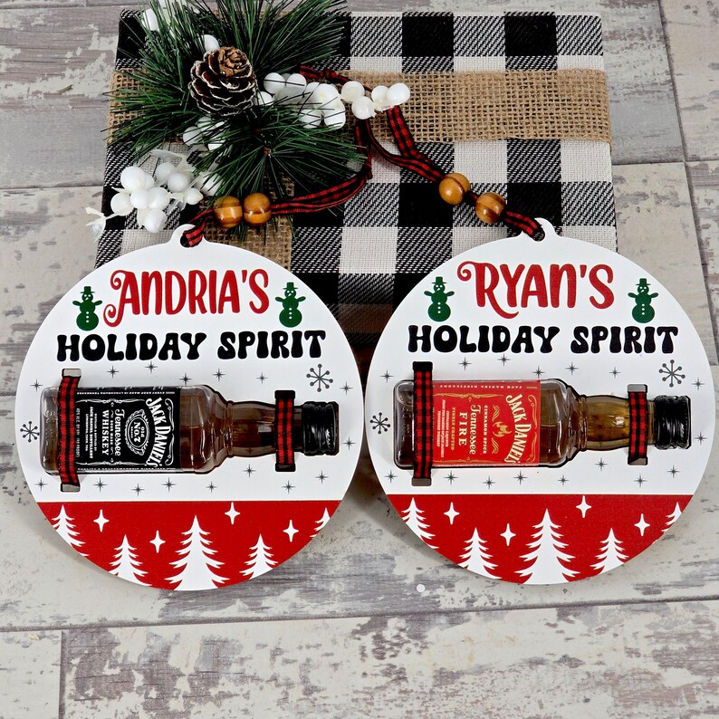 May include: Two round Christmas ornaments with the text "Andria's Holiday Spirit" and "Ryan's Holiday Spirit". Each ornament holds a miniature Jack Daniel's whiskey bottle. The ornaments have a red and white Christmas tree design.