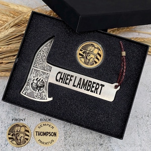 May include: A silver firefighter axe with ornate detailing and the text "CHIEF LAMBERT" is displayed in a black box. A gold coin with a firefighter graphic is also included. The box is open, revealing the contents.