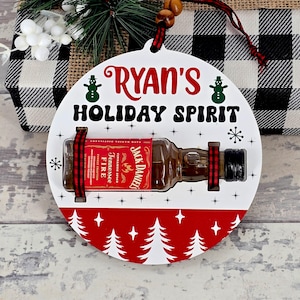 May include: A white, round Christmas ornament with the text "Ryan's Holiday Spirit" in red and green. It features a miniature Jack Daniel's Tennessee Fire bottle secured with red and black ribbon, and a red and white tree design.