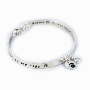 May include: Silver bangle bracelet with a paw print charm and a small clear crystal. The bracelet is engraved with the text "Always by my side" and the dates "3-17-2005".