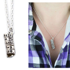 May include: A silver pendant necklace featuring a cylindrical charm. The charm is composed of stacked metal rings with engraved text. The text includes the words "esia," "Tw," "ette," "one," and the number "29." The necklace is shown against a white backdrop.