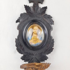 May include: A black wooden wall-mounted religious shrine with a carved cross at the top and a glass-covered oval image of Jesus Christ with a golden heart. The shrine has a carved wooden shelf below the image.