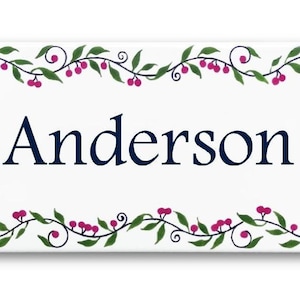 May include: White ceramic tile with a decorative border of green vines and pink berries. The name "Anderson" is written in navy blue lettering in the center of the tile.