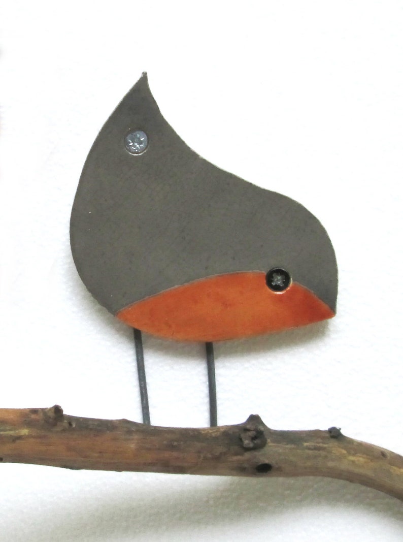 May include: A ceramic bird figurine with a gray body and an orange breast, perched on a brown branch. The bird has a simple, abstract design and is attached to the branch with metal screws.