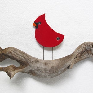 Pu&ograve; includere: Una statuetta di un cardinale rosso appollaiata su un pezzo di legno alla deriva. L'uccello ha occhi neri e un becco nero.