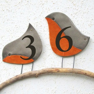 Op de afbeelding: Twee keramische vogelvormige huisnummers, een met het nummer 3 en de andere met het nummer 6, zitten op een tak. De vogels zijn grijs met oranje accenten.