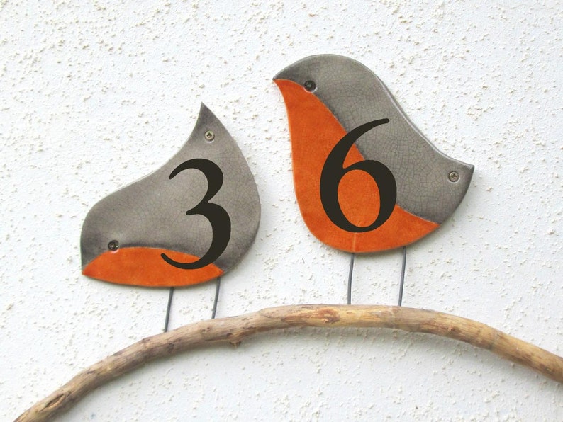 Op de afbeelding: Twee keramische vogelvormige huisnummers, een met het nummer 3 en de andere met het nummer 6, zitten op een tak. De vogels zijn grijs met oranje accenten.