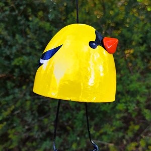 Könnte beinhalten: Ein gelber Keramikvogel mit schwarzen und weißen Markierungen und einem roten Schnabel hängt an einer schwarzen Schnur. Der Vogel hat Drahtbeine.