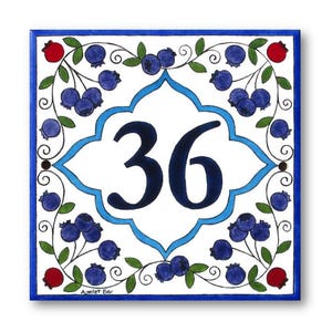 May include: A white ceramic tile with a blue and white border. The tile features a blue and white floral design with blueberries and a large number 36 in the center. The tile is signed "Ayelet Bar" in the bottom right corner.