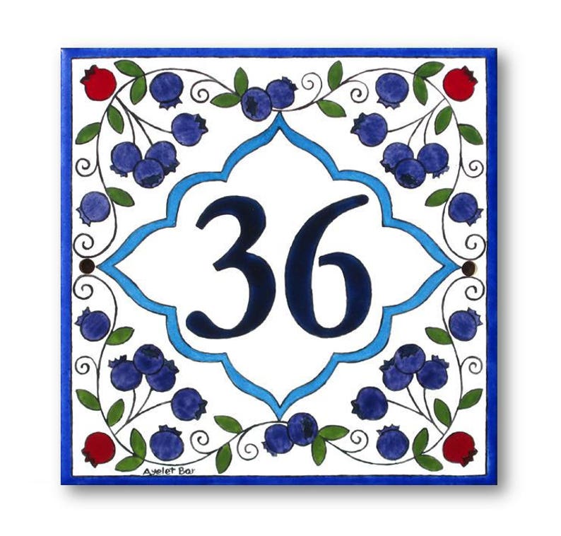 May include: A white ceramic tile with a blue and white border. The tile features a blue and white floral design with blueberries and a large number 36 in the center. The tile is signed "Ayelet Bar" in the bottom right corner.