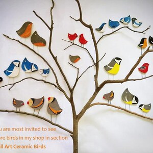 May include: A collection of colorful ceramic birds perched on a bare tree branch. The birds are painted in various shades of blue, red, yellow, and gray. The background is a plain white wall. You are most invited to see more birds in my shop in section Wall Art Ceramic Birds.