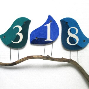 Op de afbeelding: Drie blauwe en turquoise vogelvormige ornamenten met witte cijfers 3, 1 en 8 op een tak.