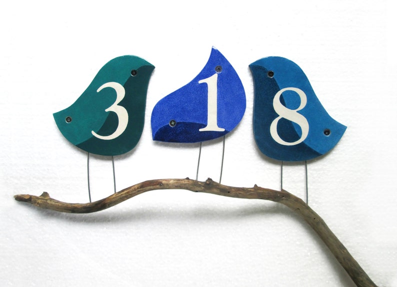 Op de afbeelding: Drie blauwe en turquoise vogelvormige ornamenten met witte cijfers 3, 1 en 8 op een tak.