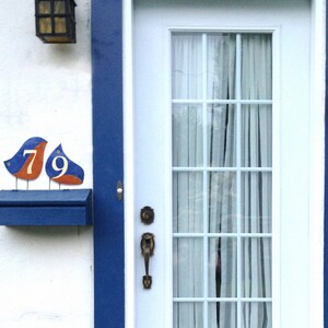 Op de afbeelding: Een blauwe brievenbus met een wit huisnummerplaatje met de nummers 79. De plaat is gevormd als twee vogels met oranje accenten. De brievenbus is gemonteerd op een witte muur met een blauwe lijst.
