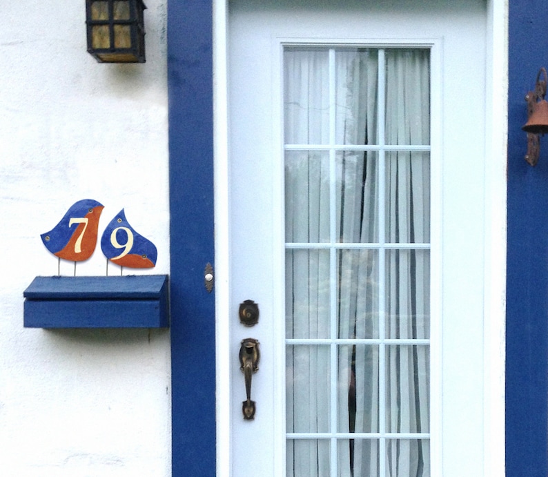 Op de afbeelding: Een blauwe brievenbus met een wit huisnummerplaatje met de nummers 79. De plaat is gevormd als twee vogels met oranje accenten. De brievenbus is gemonteerd op een witte muur met een blauwe lijst.