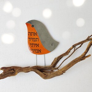 May include: A ceramic bird with orange and gray paint, perched on a driftwood branch. The bird has Hebrew writing on its body that reads "Ahava, Hamil, Ahava, Hamil".