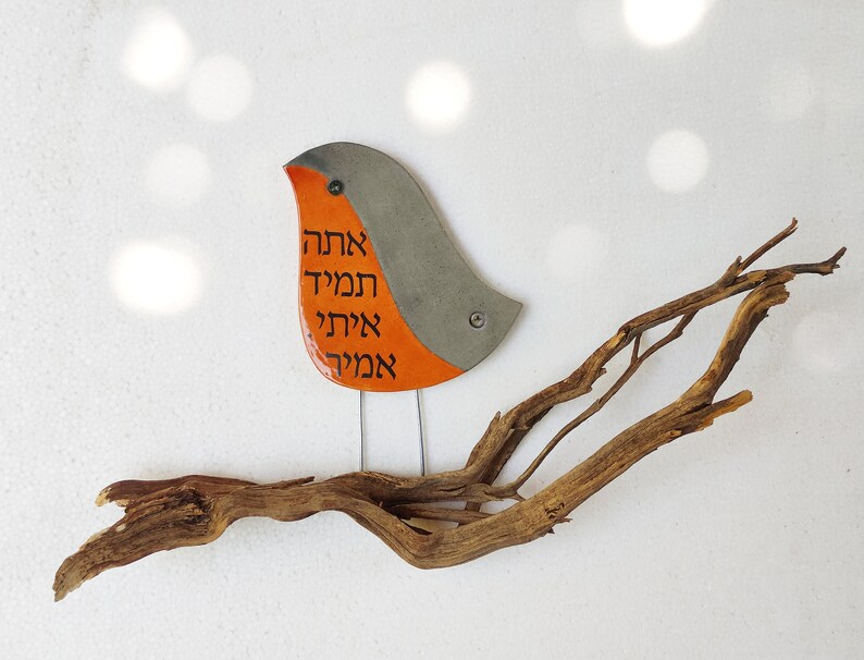 May include: A ceramic bird with orange and gray paint, perched on a driftwood branch. The bird has Hebrew writing on its body that reads "Ahava, Hamil, Ahava, Hamil".