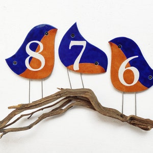 Op de afbeelding: Drie keramische vogelbeeldjes, geschilderd in blauw en oranje, elk met een wit nummer op hun borst. De nummers zijn 8, 7 en 6. De vogels zitten op een tak.