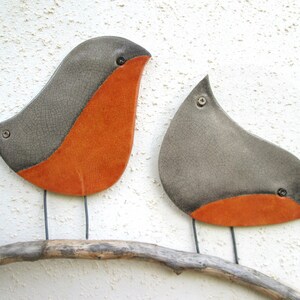 Pu&ograve; includere: Due uccelli in ceramica dipinti di grigio e arancione appollaiati su un ramo di legno galleggiante. Gli uccelli hanno una finitura smaltata craquel&eacute;.