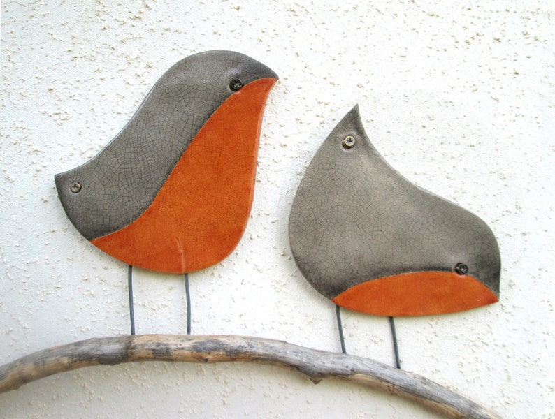Pu&ograve; includere: Due uccelli in ceramica dipinti di grigio e arancione appollaiati su un ramo di legno galleggiante. Gli uccelli hanno una finitura smaltata craquel&eacute;.
