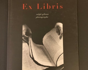 May include: A black and white photograph of a book being opened by hands. The book is open to a page with text. The title of the book is "Ex Libris" and the author is "ralph gibson photographs".