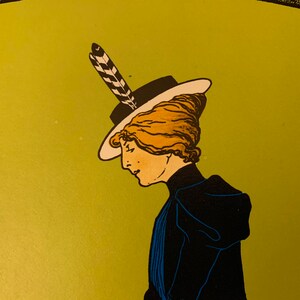 May include: A woman with blonde hair wearing a black hat with a white brim and a black and white feather. She is wearing a dark blue dress with a white collar.