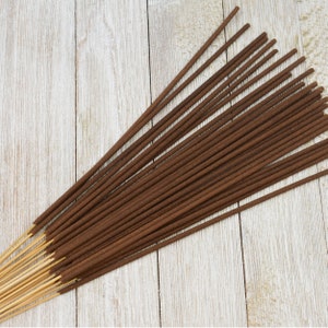 May include: A bundle of long, thin incense sticks featuring dark brown, textured aromatic material coated onto slender, light tan bamboo wooden cores. The sticks are arranged in a fan-like formation, showcasing the contrast between the dark incense coating and the exposed, smooth, light-colored wooden handles at the base.