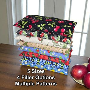 May include: A stack of seven rectangular pillows with various fruit patterns, including cherries, strawberries, and blueberries. The pillows are in different colors, such as black, white, red, blue, and tan. Text on the image reads: 5 Sizes, 4 Filler Options, Multiple Patterns.