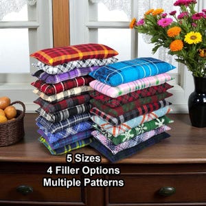 May include: Stacks of rectangular pillows in various plaid patterns and colors, including red, blue, green, and purple. The pillows are arranged on a dark wooden surface. Text on the image reads: 5 Sizes, 4 Filler Options, Multiple Patterns.