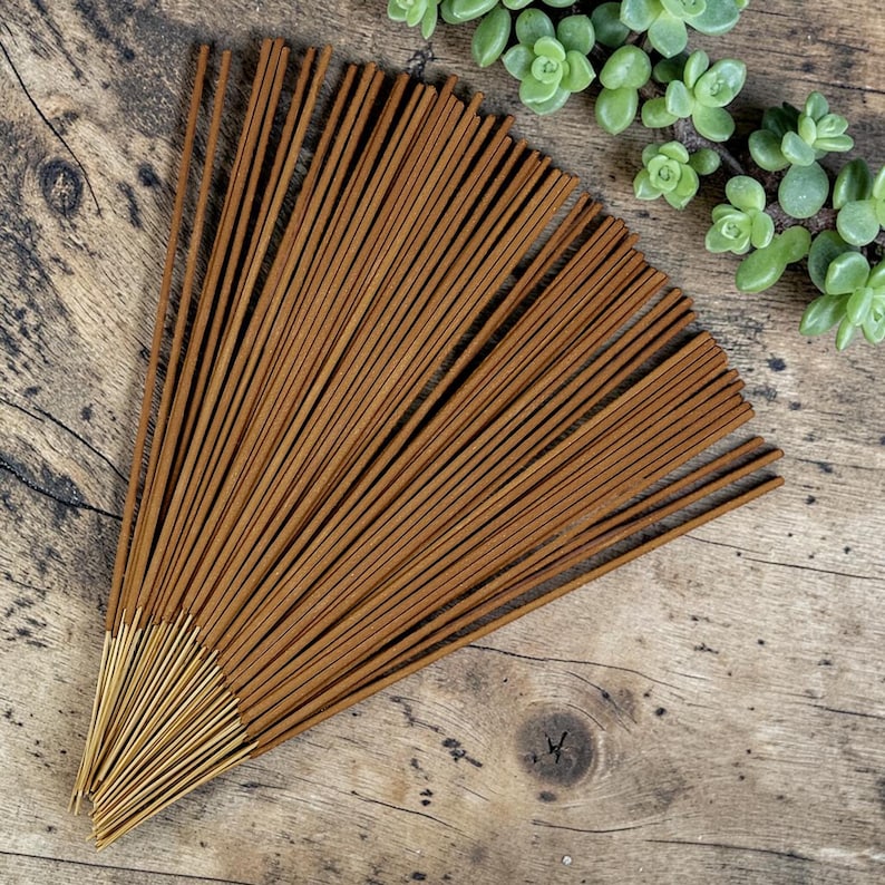May include: A collection of thin, brown incense sticks arranged in a fan shape. The sticks have a smooth, cylindrical body with a lighter, uncolored wooden base at the bottom end, fanned out to display the uniform texture and color of the incense.
