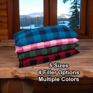 May include: A stack of four rectangular pillows featuring a buffalo plaid pattern in teal, pink, dark green, and red. The text on the image reads 5 Sizes, 4 Filler Options, Multiple Colors. The pillows are stacked horizontally on a wooden surface.