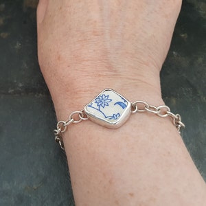 以下が含まれることがあります： 青と白の花柄の正方形のチャームが付いたシルバーチェーンブレスレットです。
