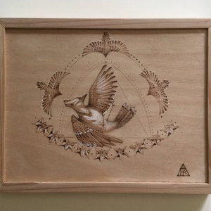 May include: A wood-burned artwork depicting a fox with wings flying in a circle surrounded by birds. The artwork is framed in a light wood frame.