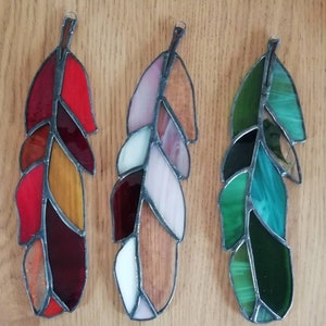 Puede incluir: Tres adornos de plumas de vidrieras en tonos rojos, rosas y verdes. Cada pluma tiene un lazo de metal para colgar. Las plumas están hechas de varios colores de vidrio, incluyendo rojo, naranja, amarillo, rosa, blanco, marrón y verde.