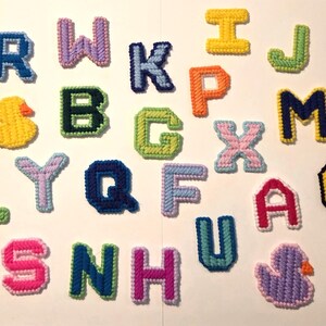 May include: A collection of colorful, handcrafted plastic canvas letters and a duck shape. The letters are in various colors, including pink, blue, green, and yellow, with white stitching. The duck is yellow with an orange beak.