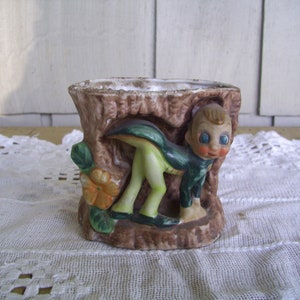 May include: A ceramic planter shaped like a tree stump with a small green and brown gnome peeking out. The gnome has large blue eyes and a brown hat.