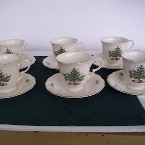 May include: Set of six cream-colored teacups and saucers. Each cup and saucer features a Christmas tree design in green, red, and gold. The rims of the cups and saucers are trimmed with a green border. A festive holiday theme.